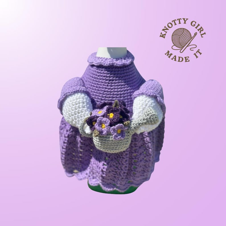 May include: A handmade crochet doll wearing a purple dress and holding a basket of flowers. The doll's sleeves are white and gray. The doll is on a light purple background. The logo "Knotty Girl Made It" is in the upper right corner.