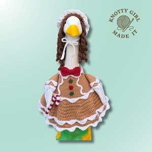May include: A crochet outfit for a decorative goose featuring a brown gingerbread-style dress with white scalloped trim, a red bow tie, and a candy cane accessory. The set includes a matching brown bonnet with white trim and the text Knotty Girl Made It.