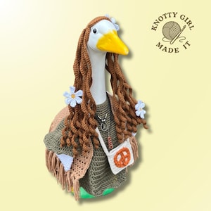 May include: A decorative goose figurine with a white head and yellow beak, adorned with brown crochet hair, blue flower accents, and a crochet outfit. The outfit includes a green top, fringed shawl, and a small white bag with a peace sign. The text "KNOTTY GIRL MADE IT" is in the upper right.