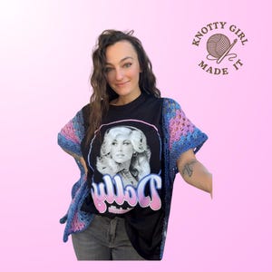 Puede incluir: Una camiseta negra con un retrato de Dolly Parton y el nombre "Dolly" en escritura rosa y azul. La camiseta se combina con un chal de ganchillo azul, rosa y morado. La imagen también incluye el texto "Knotty Girl Made It".