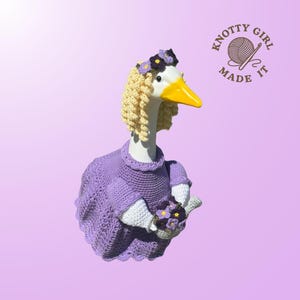 May include: A whimsical goose figurine wearing a purple crochet dress, a floral headband, and a blonde curly wig. The goose holds a bouquet of purple flowers. The image includes the text "Knotty Girl Made It."