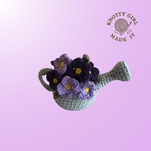 May include: A crocheted watering can filled with purple and lavender flowers with yellow centers. The watering can is light blue-green. The image includes the text "Knotty Girl Made It" in a circular logo.