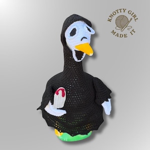 May include: A crochet goose figurine wearing a black hooded cloak and a white mask with black eye and mouth cutouts. The goose has a yellow beak and holds a white knife with a red tip. The logo Knotty Girl Made It is visible in the top right.