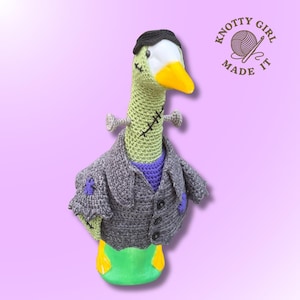 May include: A crocheted goose doll styled as a monster, featuring a light green head with black stitched scars, a black hairpiece, and a yellow beak. It wears a grey crocheted jacket with black buttons and purple patches. Text reads KNOTTY GIRL MADE IT.