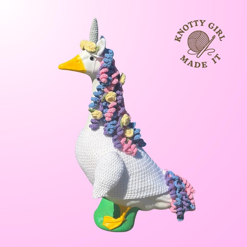 May include: A white goose figurine with a yellow beak and feet, adorned with a crocheted unicorn horn, mane, and tail in pastel colors. The goose is wearing a white crocheted sweater. The image includes the text "KNOTTY GIRL MADE IT".
