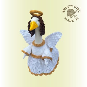 May include: A handmade crochet goose figurine dressed as an angel. The goose wears a white dress with gold trim, white wings, and a gold halo. The goose has brown hair and a yellow beak. The logo "Knotty Girl Made It" is in the upper right corner.