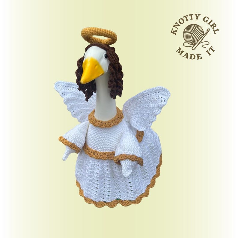 May include: A handmade crochet goose figurine dressed as an angel. The goose wears a white dress with gold trim, white wings, and a gold halo. The goose has brown hair and a yellow beak. The logo "Knotty Girl Made It" is in the upper right corner.