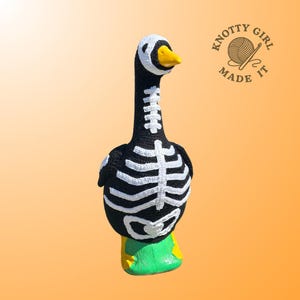 K&ouml;nnte beinhalten: Eine handgefertigte H&auml;kelgans im Halloween-Skelett-Design. Die Gans ist schwarz mit wei&szlig;en Rippen, einem wei&szlig;en Kopf und einem gelben Schnabel. Der Sockel ist gr&uuml;n und gelb. Das Logo "Knotty Girl Made It" befindet sich oben rechts.