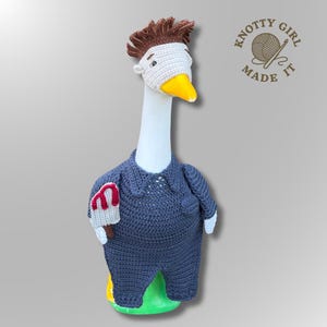 May include: A crochet goose figurine wearing a dark blue jumpsuit and a white mask with brown hair. The goose has a yellow beak and holds a small grey knife with red blood-like accents. The logo KNOTTY GIRL MADE IT is visible in the top right corner.