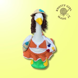 May include: A whimsical goose figurine dressed in a crocheted orange bikini, skirt, and a straw hat with a blue bow. The goose has brown curly hair and carries a beach bag and a beach ball. The image includes the text "KNOTTY GIRL MADE IT".