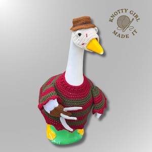 May include: A white goose figurine wearing a crocheted brown fedora and a red and dark green striped sweater. The goose has a yellow beak and a brown crocheted glove with silver-colored claws attached to its wing. The text Knotty Girl Made It is visible.