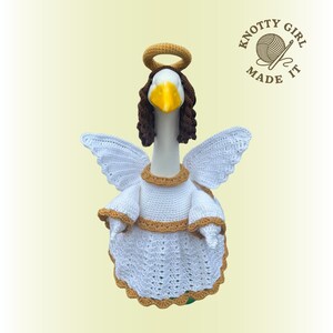 May include: A crocheted goose dressed as an angel, featuring white wings, a white dress with gold trim, and a gold halo. The goose has brown hair and a yellow beak. The text "KNOTTY GIRL MADE IT" is in the upper right corner.