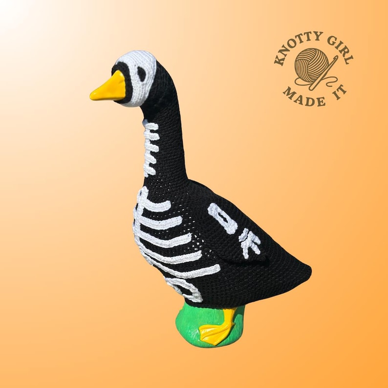 K&ouml;nnte beinhalten: Handgefertigte schwarz-wei&szlig;e H&auml;kelgans mit Skelettmuster. Die Gans hat einen gelben Schnabel und F&uuml;&szlig;e sowie einen gr&uuml;nen Sockel. Der Text "Knotty Girl Made It" ist im Bild zu sehen.