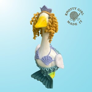 K&ouml;nnte beinhalten: Handgefertigte G&auml;nsefigur, die als Meerjungfrau gestaltet ist. Die Gans tr&auml;gt ein geh&auml;keltes, lavendelfarbenes Bikini-Oberteil, einen t&uuml;rkisfarbenen Schwanz und eine Krone. Sie hat gelocktes Haar und einen gelben Schnabel. Das Logo "Knotty Girl Made It" ist sichtbar.