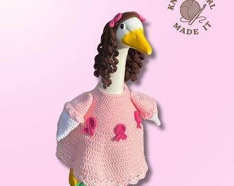 Könnte beinhalten: Eine Gänsefigur mit einem rosa Häkelkleid mit rosa Schleifen, einer braunen Lockenperücke und einer rosa Schleife. Die Gans hat einen weißen Kopf, einen gelben Schnabel und steht auf einer grün-gelben Basis. Das Logo "Knotty Girl Made It" ist sichtbar.