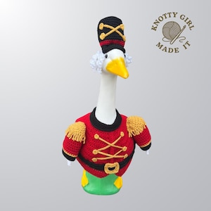 May include: A goose wearing a crocheted red and black military-style jacket, hat, and green pants. The hat is black with gold accents. The goose has a yellow beak and white hair. The image includes the text "KNOTTY GIRL MADE IT".