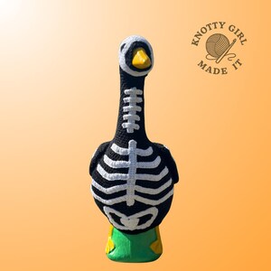 K&ouml;nnte beinhalten: Geh&auml;kelte Gans in Schwarz mit wei&szlig;en Skelett-Details. Der Schnabel ist gelb, die Basis gr&uuml;n. Der Text "Knotty Girl Made It" befindet sich in einem Kreis. Handgefertigtes Deko-Objekt.