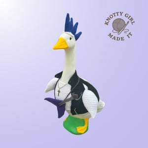 May include: A handmade crochet goose wearing a black and white jacket, a silver cross necklace, and a purple guitar. The goose has a blue mohawk and a yellow beak and feet. The image includes the text "Knotty Girl Made It".