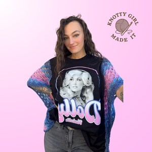 Pode incluir: Camiseta preta com um retrato de Dolly Parton e o nome "Dolly" em rosa e branco. A pessoa est&aacute; usando um xale de croch&ecirc; azul, rosa e roxo. A imagem tamb&eacute;m inclui o texto "Knotty Girl Made It".