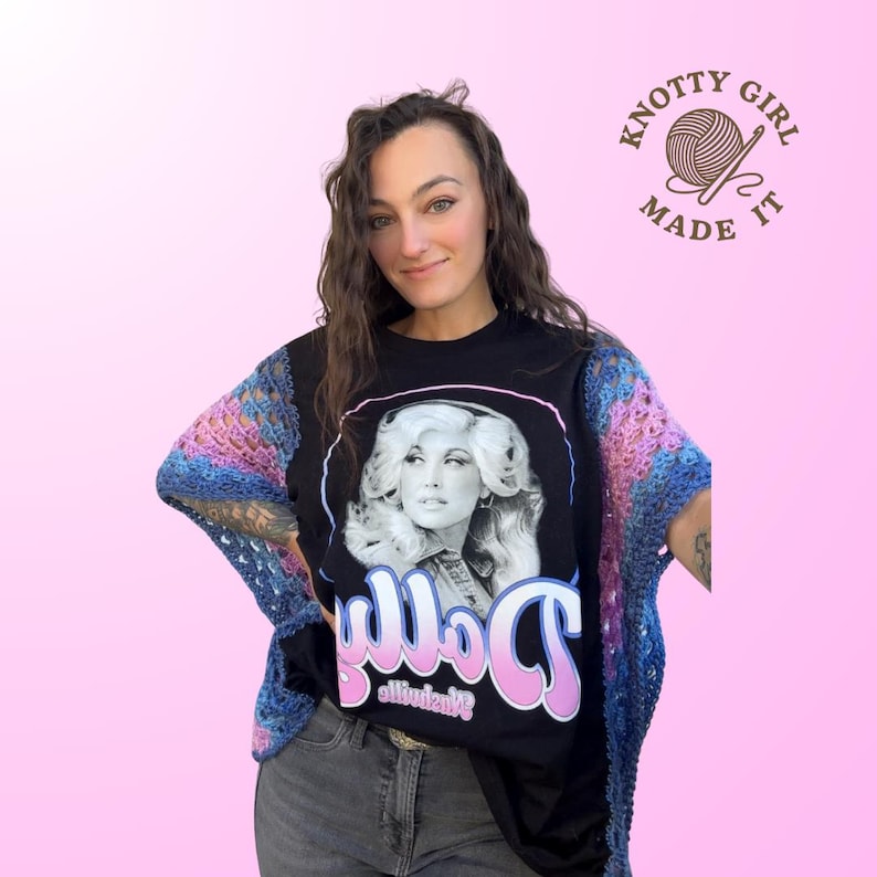 Pode incluir: Camiseta preta com um retrato de Dolly Parton e o nome "Dolly" em rosa e branco. A pessoa est&aacute; usando um xale de croch&ecirc; azul, rosa e roxo. A imagem tamb&eacute;m inclui o texto "Knotty Girl Made It".