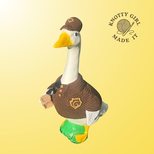Könnte beinhalten: Keramik-Gänsefigur in brauner Häkeluniform, komplett mit Mütze, Sonnenbrille und kleinem Paket. Die Gans hat einen weißen Hals, einen gelben Schnabel und grüne und gelbe Füße. Das Logo "KNOTTY GIRL MADE IT" ist oben rechts zu sehen.