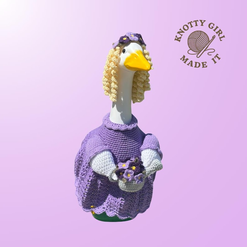 May include: A decorative goose figurine wearing a purple crocheted dress, hat, and holding a basket of flowers. The goose has blonde yarn hair and a yellow beak. The logo "Knotty Girl Made It" is in the upper right corner.