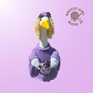 May include: A whimsical, handmade goose figurine wearing a purple crocheted dress, a floral headband, and a blonde wig. The goose holds a basket of purple flowers. The logo "Knotty Girl Made It" is visible.