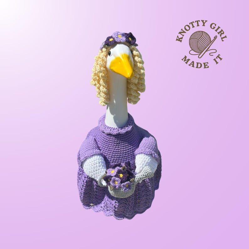 May include: A whimsical, handmade goose figurine wearing a purple crocheted dress, a floral headband, and a blonde wig. The goose holds a basket of purple flowers. The logo "Knotty Girl Made It" is visible.