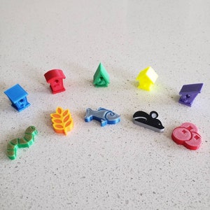 May include: A set of 10 colourful 3D printed game pieces, including a blue house, a red house, a green house, a yellow house, a purple house, a yellow wheat stalk, a blue fish, a grey mouse, a green worm, and a red berry cluster.