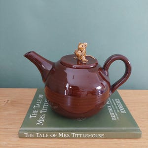 May include: A brown ceramic teapot with a mouse figurine on the lid. The teapot is sitting on a book titled "The Tale of Mrs. Tittlemouse" by Beatrix Potter.