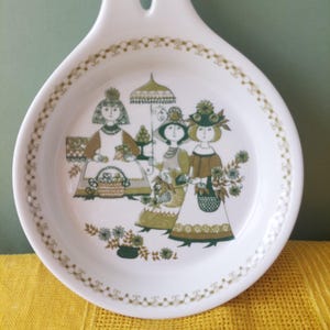 May include: A white ceramic spoon rest featuring a circular bowl and a handle with a hole. The center displays a green and brown illustration of three figures in hats, one holding an umbrella, surrounded by floral motifs and a decorative border.