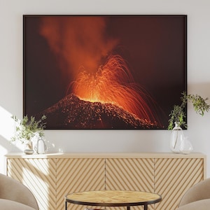 Peut inclure: Deux fauteuils beiges avec une table basse en bois et une table gigogne plus petite devant un meuble en bois avec une photographie encadrée d'un volcan en éruption avec de la lave et de la fumée.