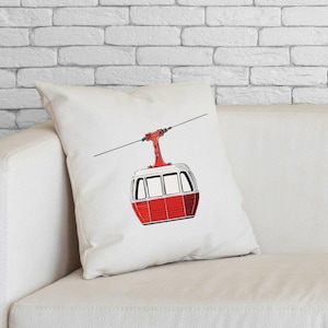 Puede incluir: Cojín cuadrado blanco con una ilustración de teleférico rojo y blanco. El teleférico está suspendido de un cable. El cojín está sobre un sofá blanco, con una pared de ladrillos blancos de fondo. El diseño es detallado.