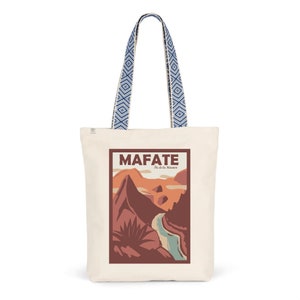 May include: A white canvas tote bag with blue and white patterned straps. The bag features a graphic design of a mountain range with a river running through it. The text "MAFATE Ile de la Réunion" is printed on the bag.