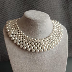Può includere: Una collana di perle a più fili esposta su un busto di gioielli grigio. La collana presenta più fili di perle bianche e rotonde, creando un effetto a cascata. Le perle variano di dimensioni, con quelle più grandi in basso e quelle più piccole in alto.