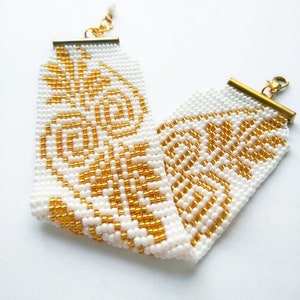Könnte beinhalten: Ein weißes und goldenes Armband mit geometrischem Muster. Das Armband ist aus kleinen, runden Perlen gefertigt und hat einen Verschluss.
