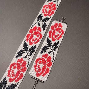 Peut inclure: Deux bracelets en perles avec un fond blanc et des motifs floraux rouges et noirs. Les bracelets sont reliés par une chaîne argentée.