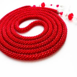 May include: A coiled, vibrant red beaded necklace. The necklace is made of small, closely-knit beads, forming a thick, rope-like structure. A delicate strand of red and clear beads extends from the main coil, adding a contrasting element. The background is plain white.