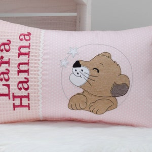 Könnte beinhalten: Ein rosa und weißes Kissen mit einem rosa Punkte-Muster. Das Kissen hat einen weißen gestickten Rand und den Namen "Lara Hanna" in rosa gestickt. Das Kissen zeigt außerdem ein gesticktes braunes Löwenjunges mit einer rosa Nase und einem rosa Hintergrund.