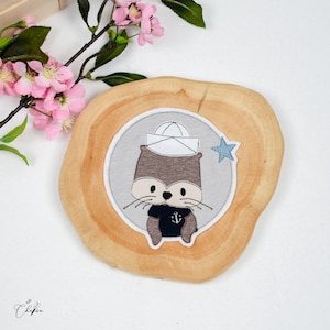 Könnte beinhalten: Eine braune und graue Filzapplikation einer Otter, die einen weißen Matrosenhut und ein blaues Hemd mit einem Anker trägt. Die Applikation befindet sich auf einer runden Holzplatte mit einem hellblauen Stern.