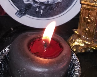 May include: A lit, dark gray candle with a red wax center sits in a decorative silver dish. Behind the candle is a circular image of a witch flying on a broomstick. A small, gold-colored decorative object is also visible.