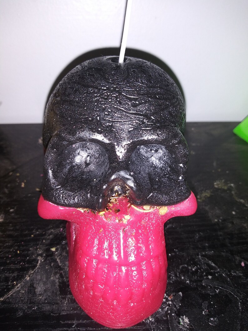 May include: A black and red skull-shaped candle with a wick. The top of the skull is black, and the jaw is red. The candle is on a dark surface.