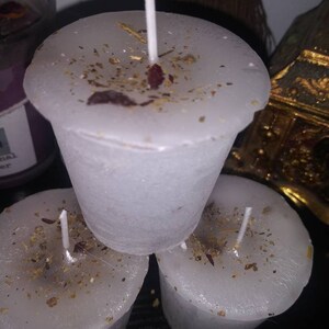 May include: Three white votive candles with a sprinkling of dried herbs and petals. Each candle has a white wick. The candles are arranged in a small stack, with one candle on top of the other two.