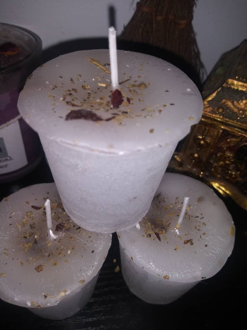 May include: Three white votive candles with a sprinkling of dried herbs and petals. Each candle has a white wick. The candles are arranged in a small stack, with one candle on top of the other two.