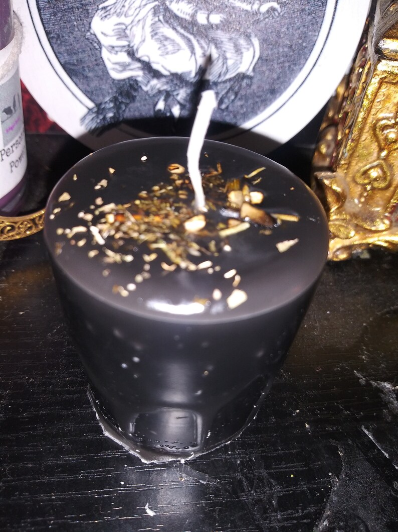 May include: A black candle with a white wick, decorated with dried herbs and gold flakes. The cylindrical candle is on a dark surface. Decorative items are visible in the background.