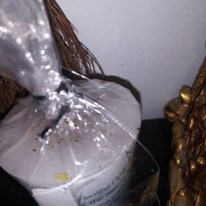 May include: A white candle wrapped in clear plastic, with text visible on the label. The text reads "Healing Spell" and other words. A bundle of brown twigs and a gold-colored decorative object are also in the image.