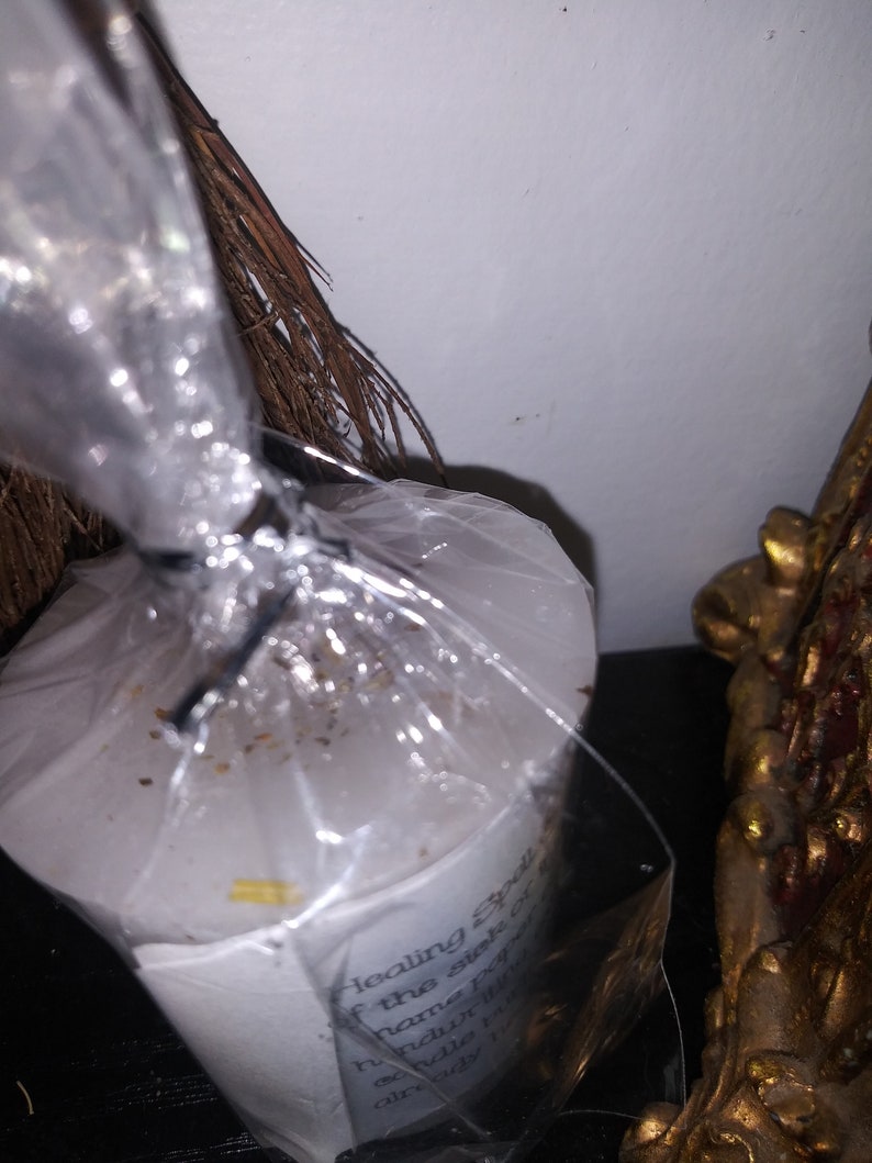 May include: A white candle wrapped in clear plastic, with text visible on the label. The text reads "Healing Spell" and other words. A bundle of brown twigs and a gold-colored decorative object are also in the image.