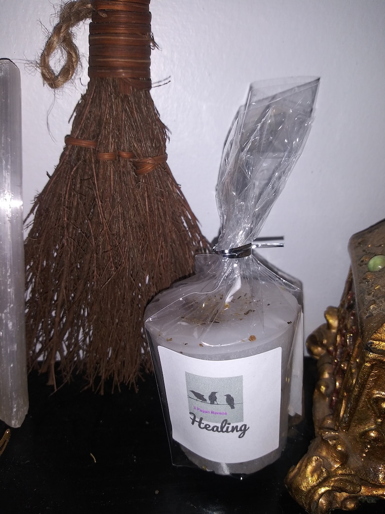 May include: A white candle with the word "Healing" on a label, packaged in clear cellophane. The label features three raven silhouettes. A small brown broom and a white crystal are in the background.