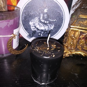 May include: A black votive candle with a white wick, decorated with dried herbs and spices. A round coaster with a black and white illustration of a witch flying on a broomstick is behind the candle. A purple candle and a gold decorative box are also visible.