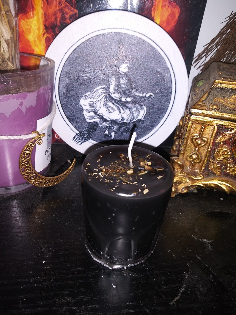 May include: A black votive candle with a white wick, decorated with dried herbs and spices. A round coaster with a black and white illustration of a witch flying on a broomstick is behind the candle. A purple candle and a gold decorative box are also visible.